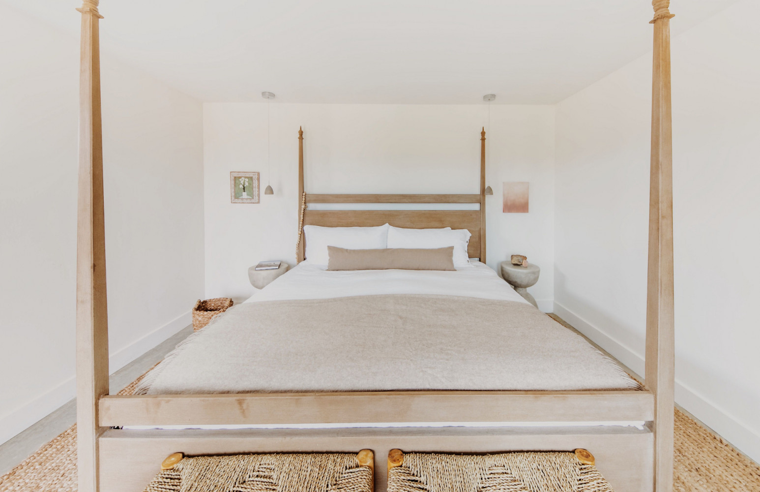 A neatly made bed with a light wood four-poster frame stands in a corner of a white-walled room with small nightstands and a woven basket at the foot.