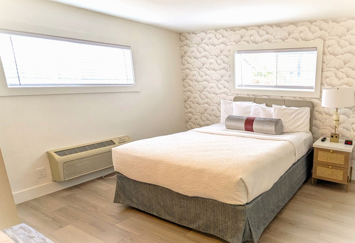 A neatly made bed with white linens and a gray bed skirt in a room with light-colored wood floors, white walls, and a patterned wallpaper headboard.