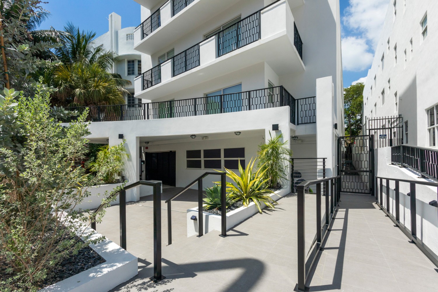 A white, multi-story building with balconies and black railings, surrounded by plants and a gate.