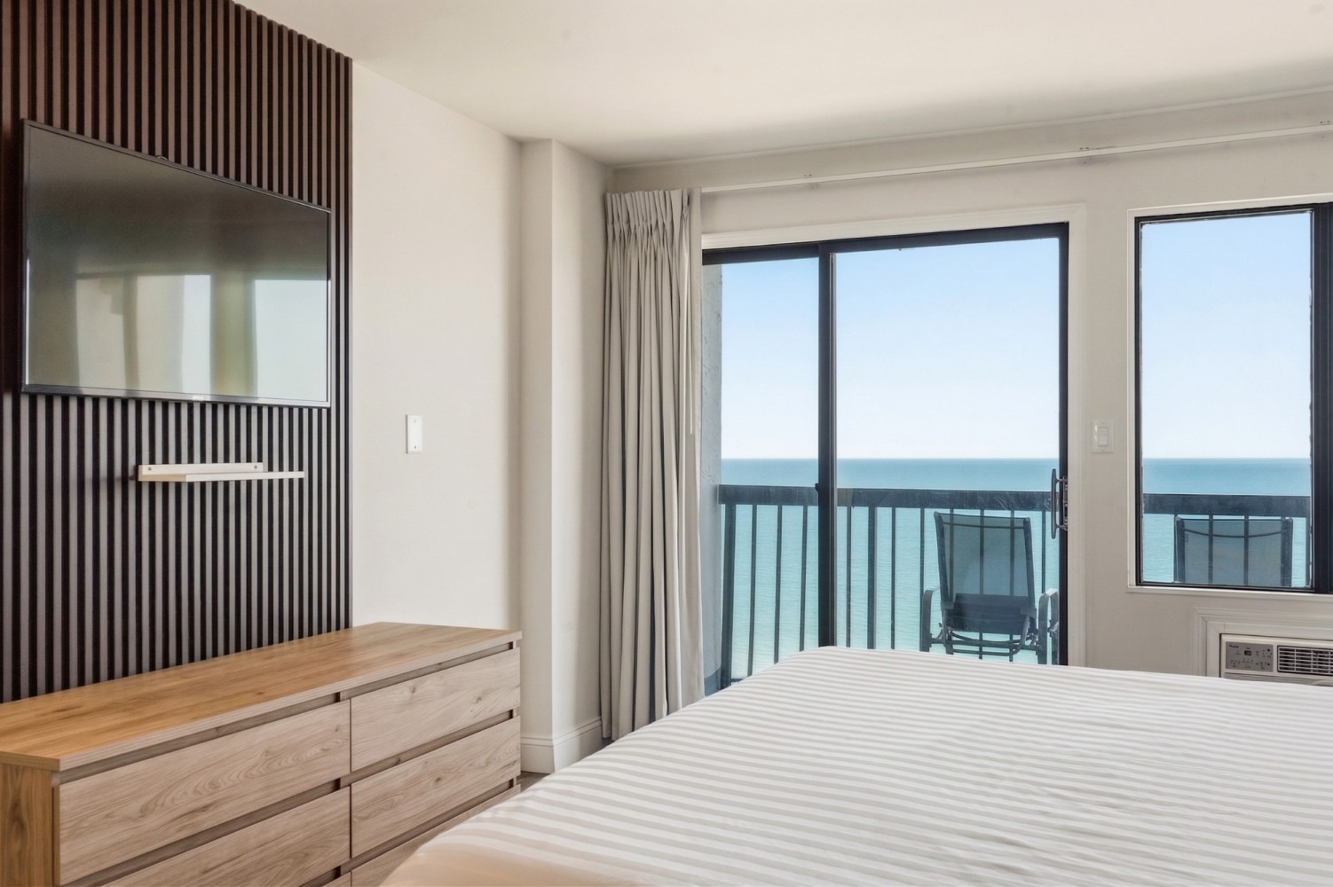 A hotel room with a large bed, wooden dresser, flat-screen TV, and balcony access with an ocean view.
