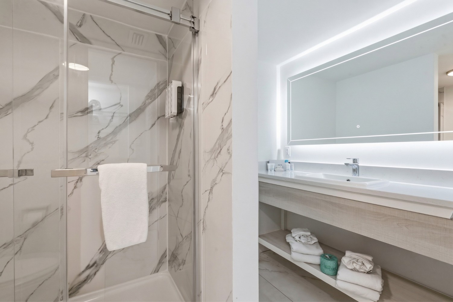 A modern bathroom with a glass-enclosed shower, marble walls, and a lit mirror above a floating sink.