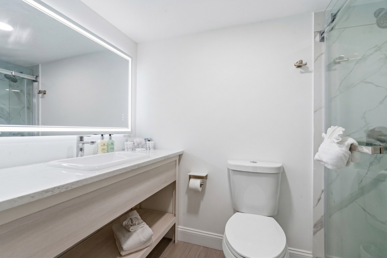 A clean and modern bathroom with a large mirror, marble-like tiles, and a glass-enclosed shower.