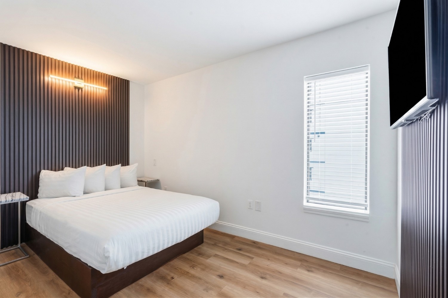 A hotel room with a large bed, a window with blinds, and a flat-screen TV mounted on the wall.