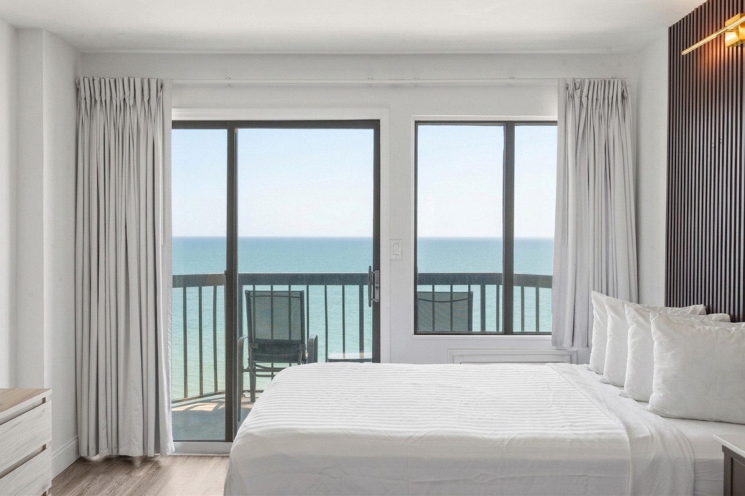 A neatly made bed with white linens and pillows in a room with a large window and sliding glass doors leading to a balcony with an ocean view.