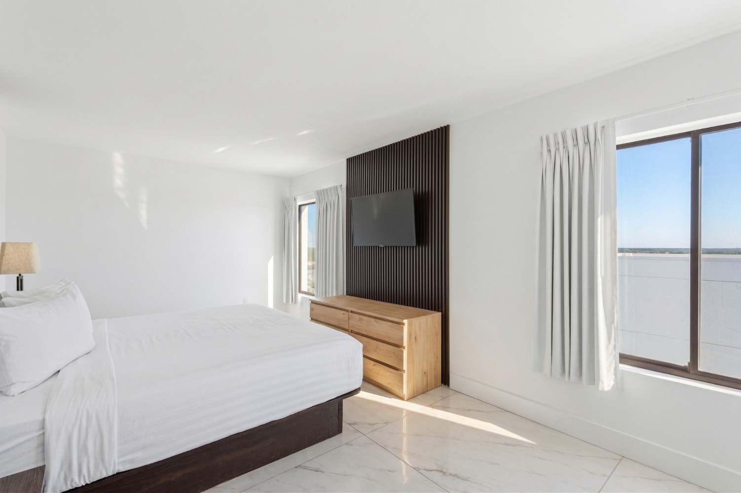 A wide view of a clean and modern guest room with a large bed, wooden dresser, mounted TV, and windows with a view.